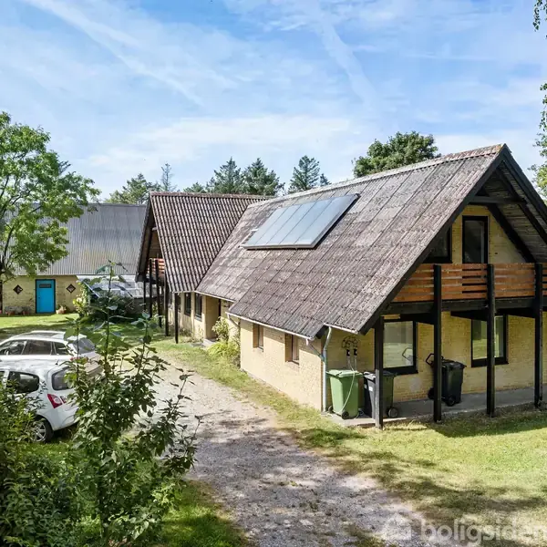 Et to-etagers hus med brunlige tegl, solpanel på taget og balkon. Flere biler er parkeret på grusvejen i en frodig, skovlignende have.