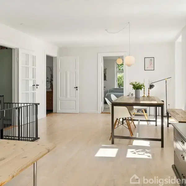 En stue med lyse trægulve indeholder en brændeovn i venstre hjørne. Et spisebord står centralt med en lampe over. I baggrunden ses en åben dør til et andet rum.