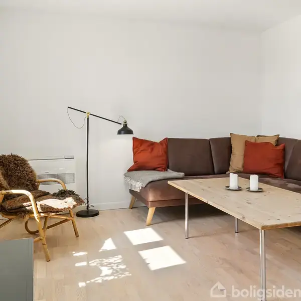 En brun hjørnesofa med farverige puder står i en lys stue. Ved siden af er en gulvlampe og et træbord med to stearinlys. En stol med fåreskind er ved vinduet.