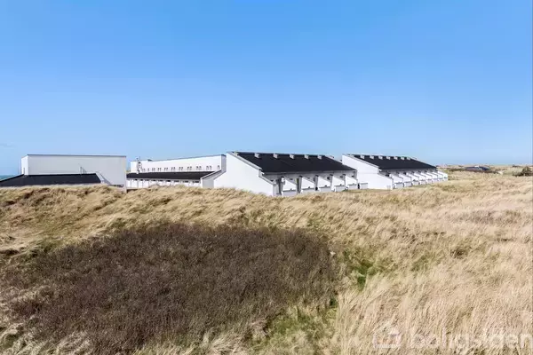 Bygninger med flade tage står stille i en række med solpaneler, omkranset af en græsbevokset klitlandskab under en klar blå himmel.