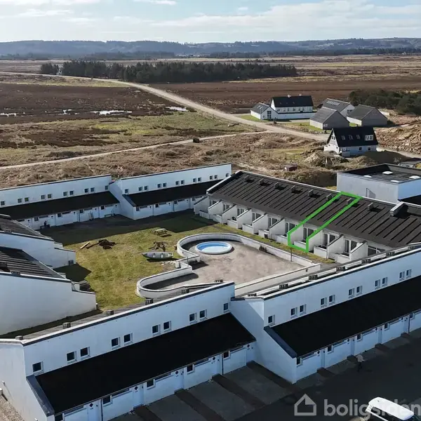 Et hvidt, L-formet boligkompleks omkranser en central gårdhave med en lille legeplads og sandkasse. Omkringliggende landskab består af åbne marker og en vej, der fører til flere huse.