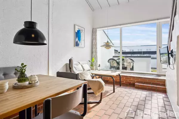 Et moderne, lyst rum med spisebord og lamper, afslappende stole ved et stort vindue. Udsigt til en balkon med stole. Brune klinker og hvidmalede vægge præger indretningen.