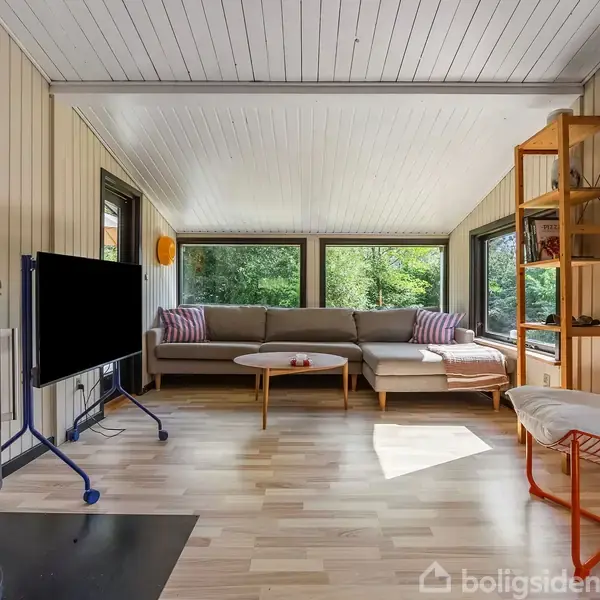 Brændeovn er i gang, placeret i en stue. En grå sofa står foran store vinduer, udsigt til natur. Et sofabord og et TV på gulvet kompletterer rummet.