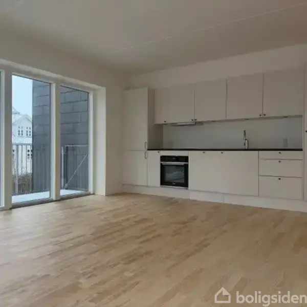 En rummelig og lys stue med trægulv. Et moderne, hvidt køkken langs muren indeholder en ovn og vask. Store vinduer til venstre giver udsigt til bygninger udenfor.