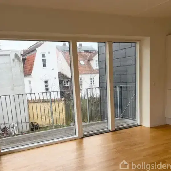 Store glasdøre åbner ud til en altan, med udsigt over nabohuse med røde tage. Indenfor er der et lyst trægulv og et køkkenhjørne til højre.