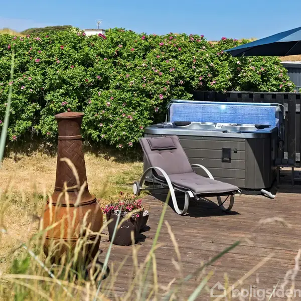 En terrasse med en liggestol ved siden af et udendørs spabad. En stor lerovn står i nærheden, og en parasol giver skygge. Omkring terrassen er der frodige buske.