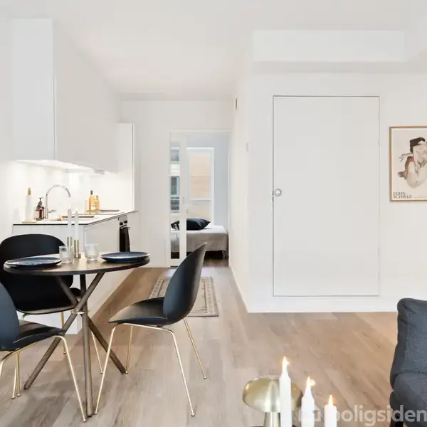 Et moderne køkken-alrum med et rundt spisebord og to sorte stole. En sofa står til højre, og en plakat hænger på væggen. Lys indstrømmer fra åbne døre.