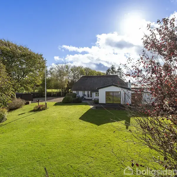 Et hvidt hus med stråtag står i en grøn have badet i sollys, omgivet af træer og blå himmel med lette skyer.