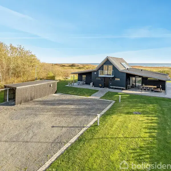Et sortmalet hus med skråtag står i et åbent område omgivet af træer. Foran huset er en grusbelagt indkørsel og en græsplæne. Solrig himmel ses i baggrunden.