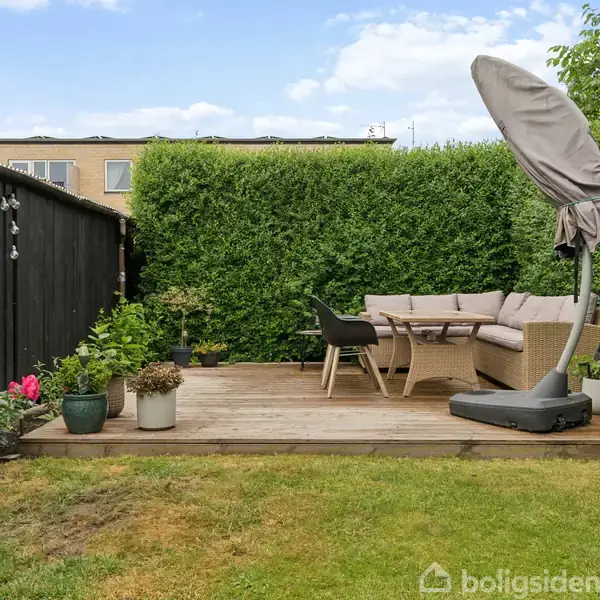 Terrasse med bord og stole i en have. En lukket parasol står ved siden af sofaen. Omkring terrassen er der grønne buske og en sort træhegn med plantede blomster.