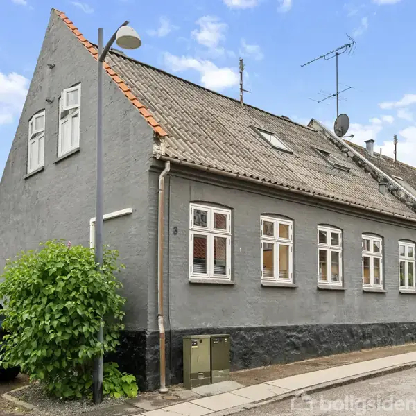 Gråt murstenshus med skråt tegltag, placeret i en byvej. En parkeret bil og en gadelygte ses ved siden af huset.