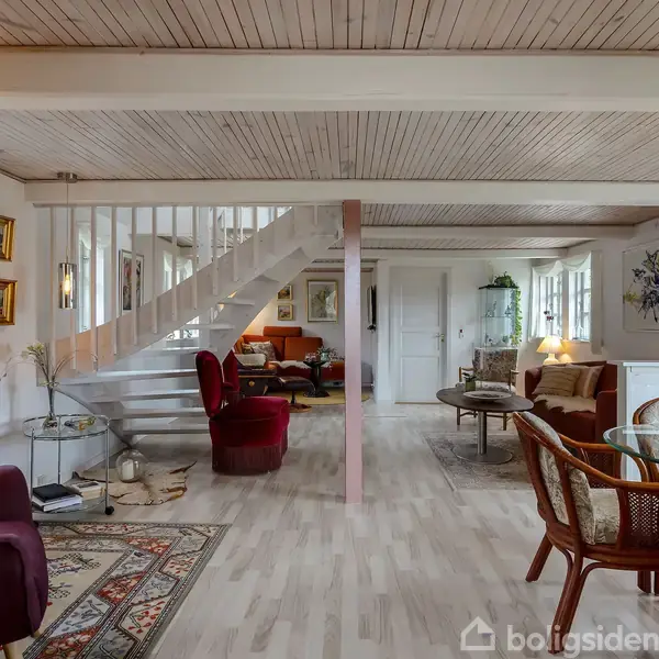 Stue med sofaer og stole placeret rundt et sofabord, en trappe op til første sal. Lyse vægge, dekorative malerier og store vinduer skaber en lys og indbydende atmosfære.