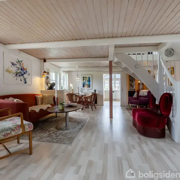 Stue med lyse trægulve har sofa, lænestole og spisebord, omgivet af vinduer. En trappe leder til en øvre etage, og der hænger kunst på væggen.