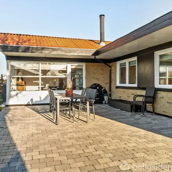 Terrasse med flisebelægning og havemøbler, herunder bord og stole, foran gulstenshus med store vinduer. Græsplæne og buske i baggrunden, med klar himmel og sollys.