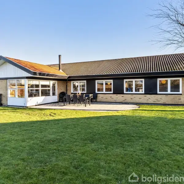 En etplans murstenshus har en terrasse med havemøbler foran. Huset ligger i en velplejet have med kortklippet græs og et træ i baggrunden under en klar blå himmel.