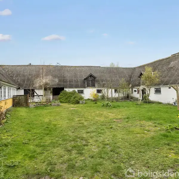 En gårdsplads omgivet af gamle landbygninger med stråtaget. Den venstre bygning er gul med hvide vinduer. Græsset dækker jorden, og der er et par planter og træer.