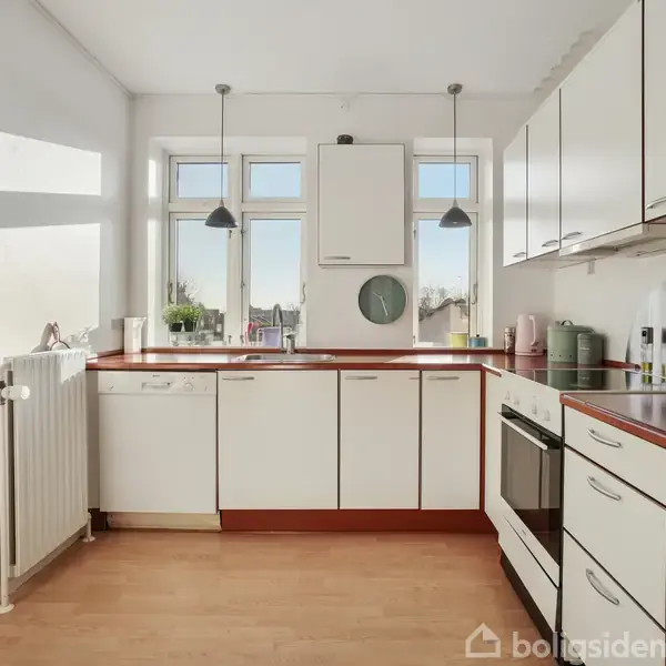 Hvidt køkken med rød bordplade, ovn og vask. Der er hængende lamper over vinduet, og køkkenet er lyst af naturligt dagslys. Radiator ved siden af en åben dør.