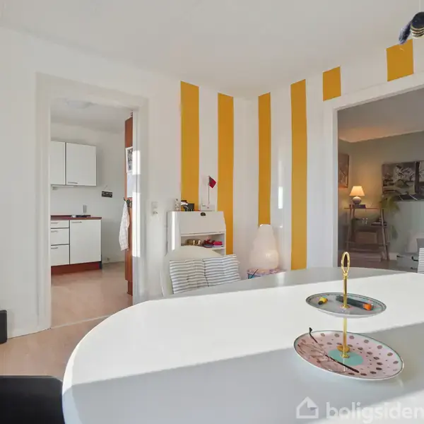 Et rundt bord står i et rum med gule og hvide stribede vægge. En turkis rullebord står ved siden af. Køkkenet ses gennem en åben dør.