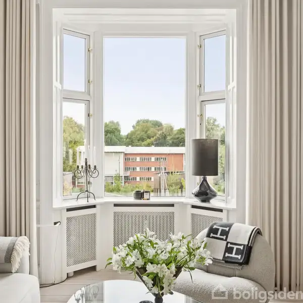 Et lyst, elegant stuehjørne med en stor karnapvindue viser udsigt til en rød bygning og træer udenfor. En sofa, bord med blomster og lampe skaber hyggelig atmosfære.
