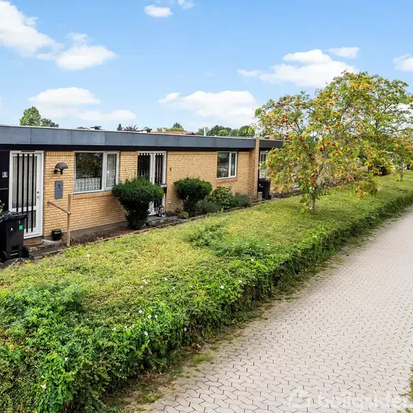 En murstensbungalow med sort tag står ved siden af en flisebelagt sti. Omkring huset er der grønne buske og træer. Baggrunden viser flere huse og en blå himmel med skyer.
