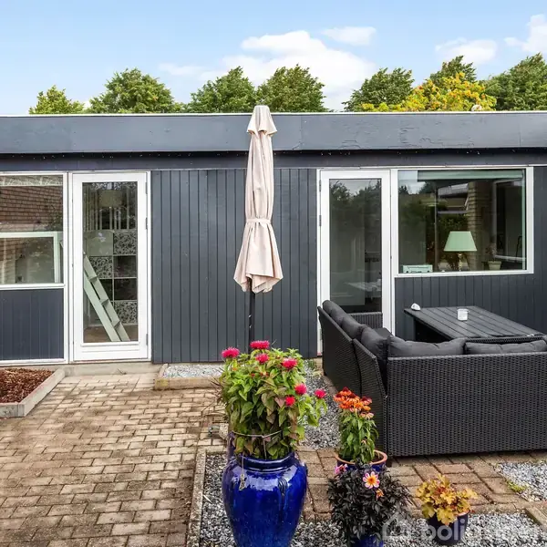 En moderne terrasse har havemøbler, en parasol og blomster i en blå vase. Huset omkring er gråt med store vinduer. Baggrund har grønne træer og blå himmel.
