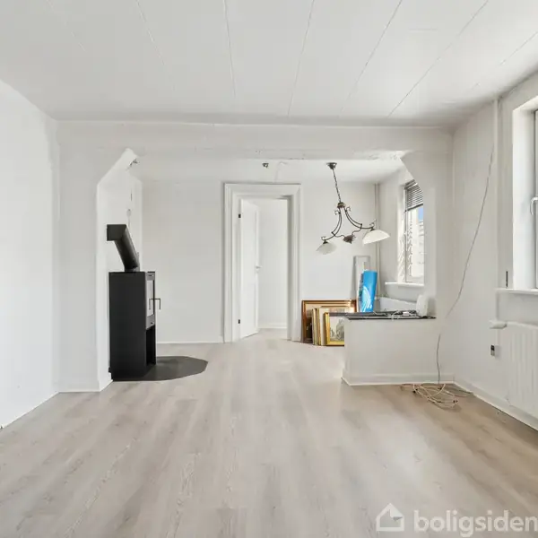 En lys stue har en brændeovn til højre. To vinduer slipper lys ind. Der er en lysekrone og et åbent rum i baggrunden. Væggene er hvide, gulvet lyst.