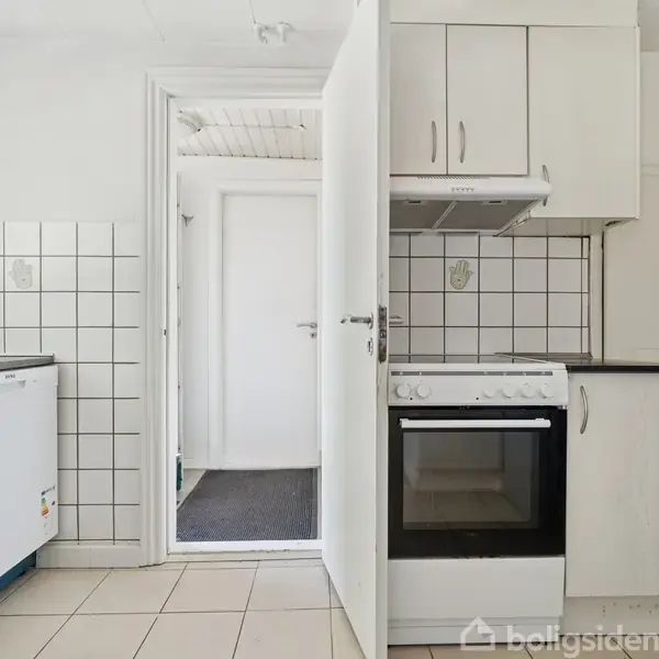 Komfur med emhætte og skabe i et lille køkken. Opvaskemaskine til venstre. Flisebelagte vægge og lyse fliser på gulvet. Åben dør fører til en lys gang.