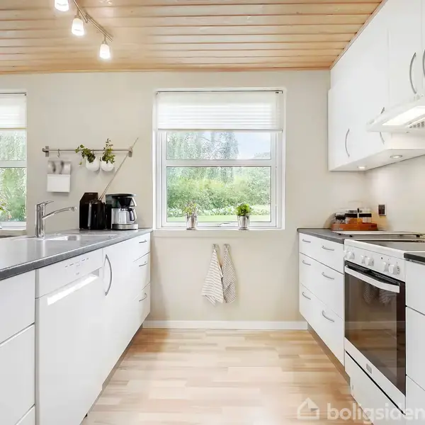 Et moderne køkken med hvide skabe og sorte bordplader. En kaffemaskine og køkkenredskaber står på bordet. To store vinduer slipper masser af lys ind. Trægulv og loft.