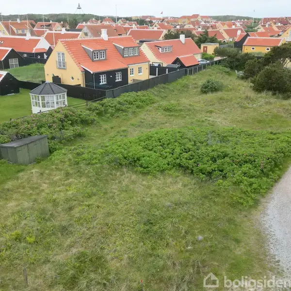En grusvej snor sig forbi grønne marker foran gule huse med røde tage. En lille bygning står tæt ved vejen, og flere huse ses i baggrunden.