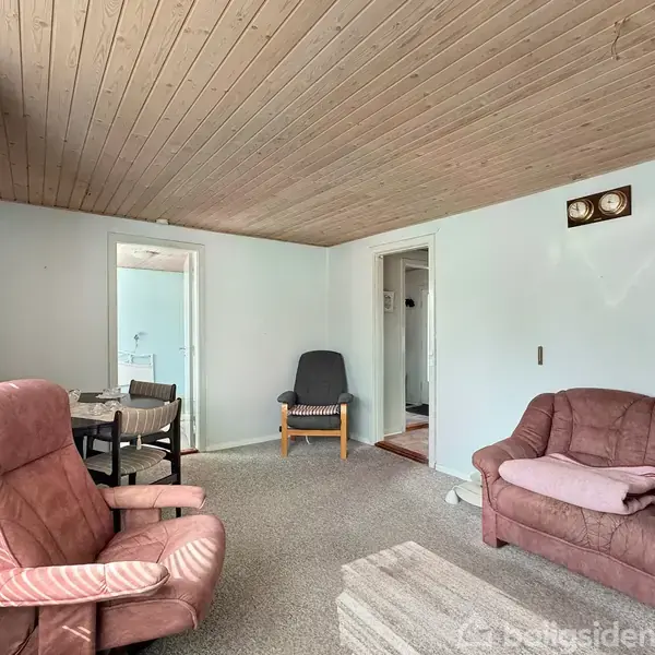 Stue med pink sofa og matchende lænestol står på et tæppebelagt gulv. En spiseplads med borde og stole ses i baggrunden. Vægge og loft er træbeklædte. Ingen tekst.
