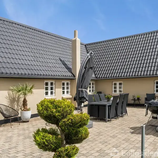Gårdhave med møbleret terrasse, herunder et udendørs spisebord, parasol og grill; omgivet af beige vægge og sort tegltag. Potteplanter og geometrisk busk står langs den brostensbelagte gårdsplads.