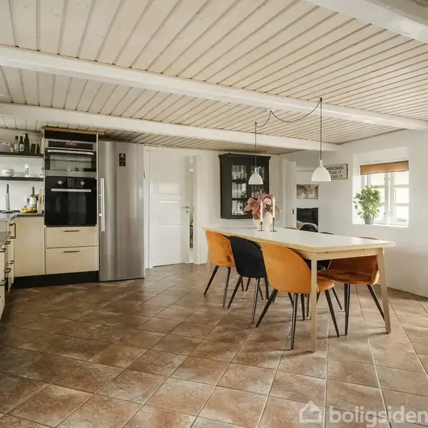 Et moderne køkken med beige skabe og sorte håndtag har en bardisk med hvide lamper. Et spisebord med seks orange stole står centralt. Rummet har lyse fliser og træloft.