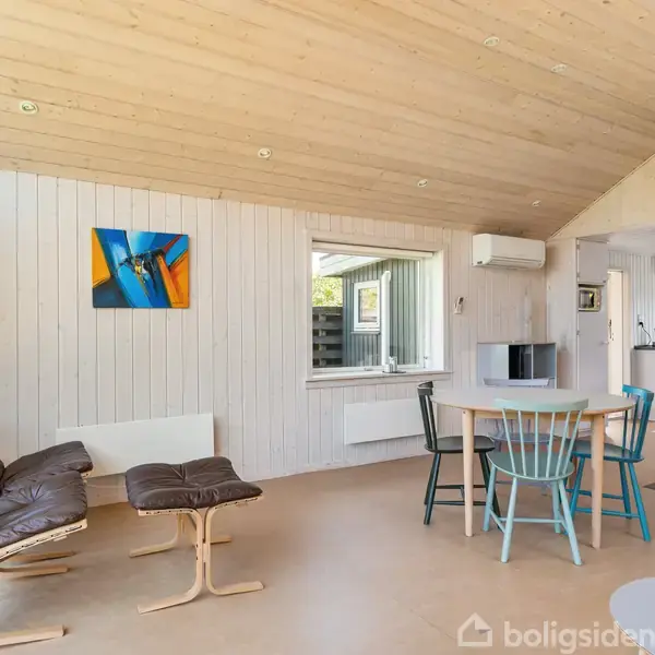 En stue med en brun lænestol og skammel, der står ved et rundt spisebord med forskellige stole. En moderne brændeovn står i hjørnet. Væggene er lyse med kunst.