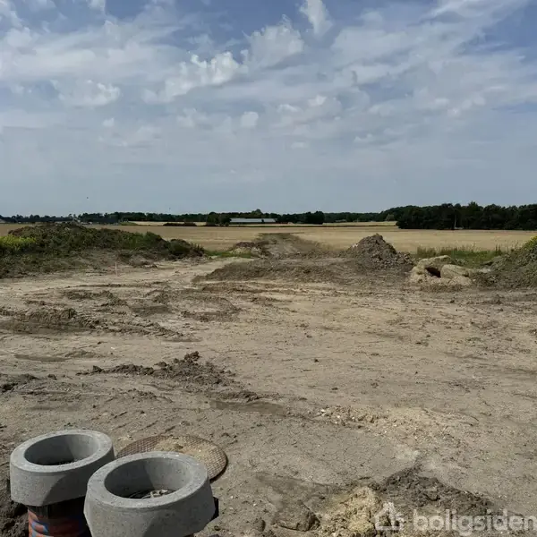 Jordområde med udgravninger, stille under skyet himmel, omgivet af marker og træer i det fjerne. Ingen synlig aktivitet eller bygninger i nærheden.