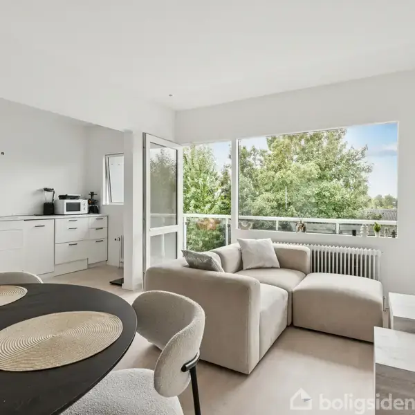 En lys stue med en beige sofa hjørne mod et stort vindue, der viser træer udenfor. Til venstre ses et hvidt køkken med komfur og mikrobølgeovn.