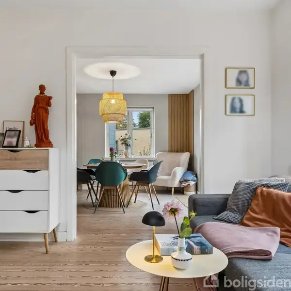 En lys stue med en grå sofa, pyntepuder, og et lille rundt bord. Fra stuen fører en åbning til en spisestue med blå stole rundt om et bord.