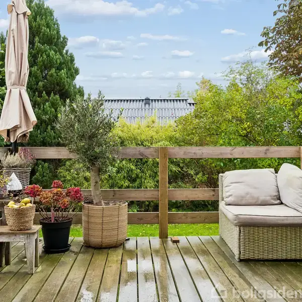Sofa står stille på en træterrasse med udsigt til en frodig have. Ved siden af er der potteplanter, en parasol og himlen er let skyet.