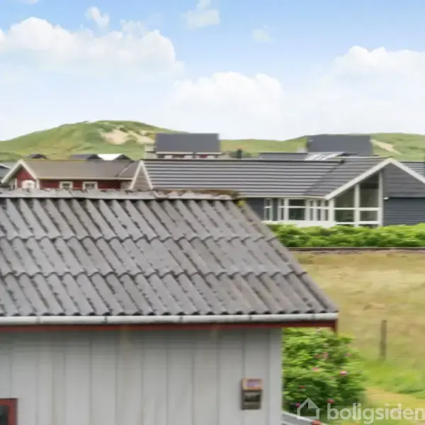 Et gråt tag på et hus med udsigt til grønne marker og bakker i baggrunden. Flere huse ses i horisonten under en blå himmel med skyer.