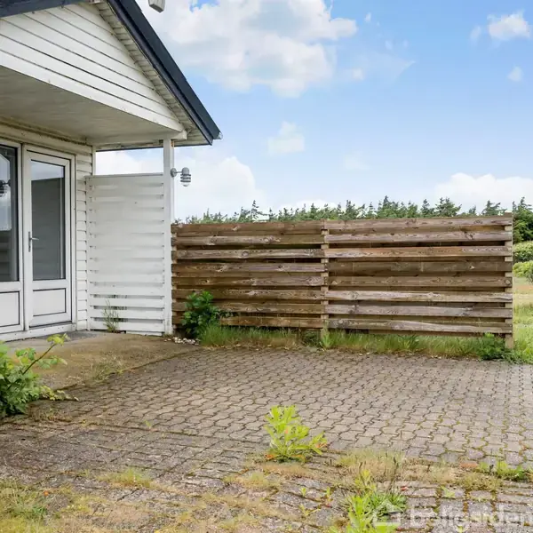Husindgang med nummer 44, omgivet af en flisebelagt terrasse og en træhegn beliggende i et grønt område med buske og en klar blå himmel i baggrunden.