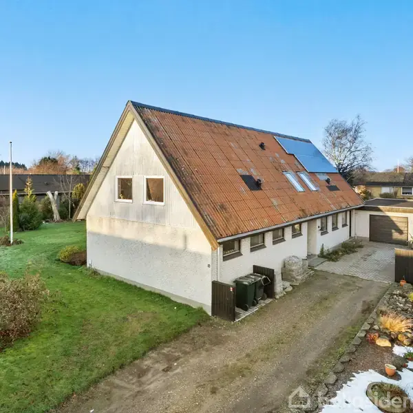 Et hvidt hus med rødt tegltag og solpaneler står omgivet af en grøn have. Til højre ses en grusvej og en indhegnet have.