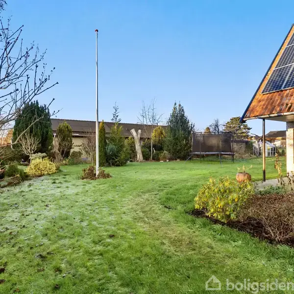 Hus med solpaneler på taget står i en have med grønt græs og buske. En flagstang står i det åbne område, omgivet af træer under en blå himmel.