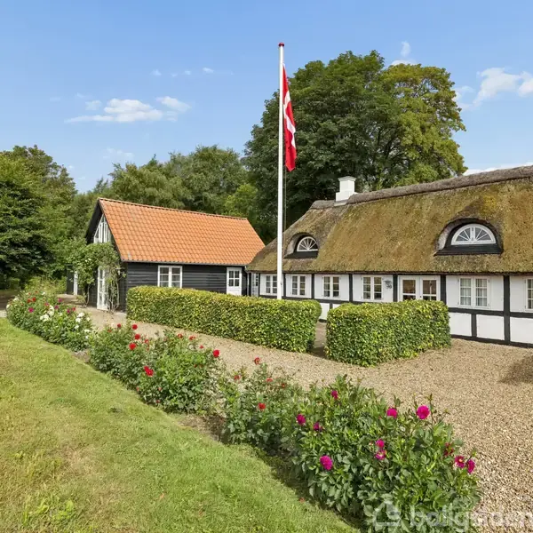 Et stråtækt bindingsværkshus står stille omgivet af en grussti, velplejede buske og blomster. Det danske flag vajer på en stang, mens træer danner baggrundens grønne kulisse.