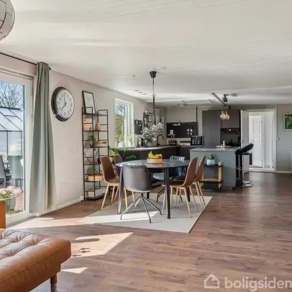 Stue med spisebord og stole, sofa ved siden af store vinduer. Køkken i baggrunden med moderne indretning. Væggene prydet med ur og dekorationer, træplankegulv.