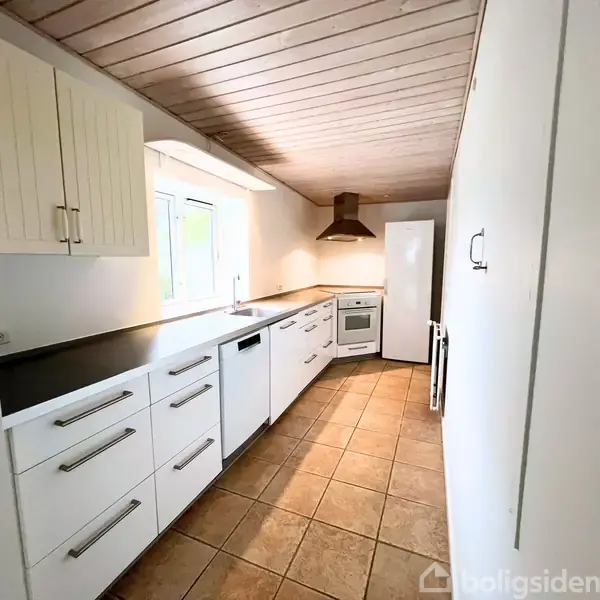 Et smalt køkken med hvide skabe og sort bordplade. En ovn og ventilator er placeret i slutningen af rummet. Flisegulvet skaber en lys og moderne atmosfære.