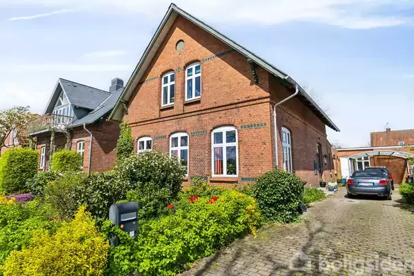 Murstenshus med hvide vinduer står i en frodig have med farverige blomster. En bil er parkeret på grusvejen ved siden af huset. Det er solskinsvejr.