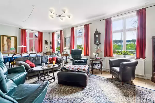 Stue med sorte lædermøbler og en stor gulvlampe i et rum med mange vinduer, røde gardiner og udsigt til en have. Orientalsk tæppe dækker gulvet.