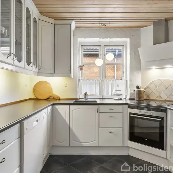 Hvidt køkken med sorte bordplader. Indbygningsovn og keramisk kogeplade er integreret i hjørnet. Vintersina vindue med gardin over vask. Skærebræt, planter og krydderier pynter.