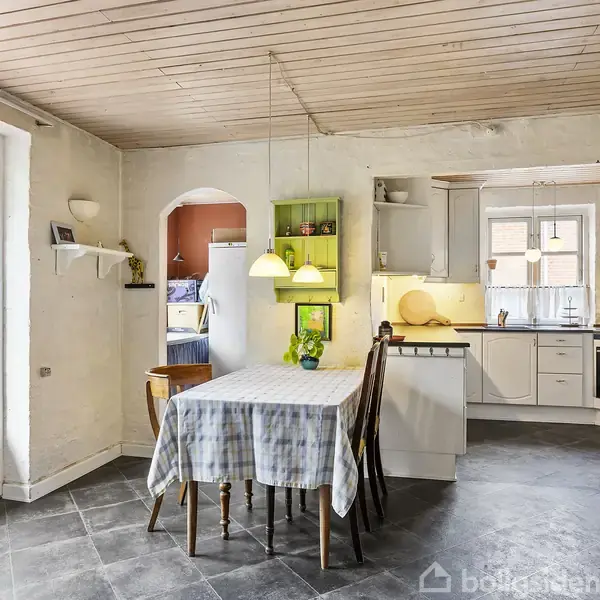 Spisestue med træbord dækket af ternet dug, omgivet af stole. Lamper hænger over bordet. I baggrunden ses et åbent køkken med hvide skabe og apparater.