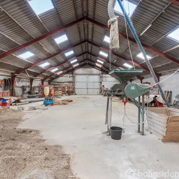 En stor lade med metalramme indeholder landbrugsudstyr, inklusive en tragtmaskine. Der er værktøj og materialer spredt langs væggene, og gulvet er delvist dækket af halm.