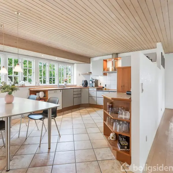 Et køkken-alrum med spisebord og stole midt i rummet. Bordet har en vase med blomster, og køkkenet er i baggrunden med hvide skabe. Store vinduer giver udsigt til grønne træer.